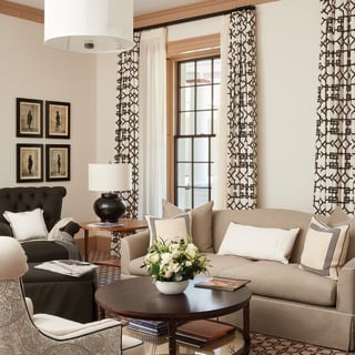 A stylish airy living area with a beige sofa and black upholstered armchair, with cream and monotone print drapes and carpet.