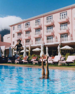 Action shot as man cannon-jumps into the pool where a swimmer performs an underwater handstand, overlooked by the pink hotel.