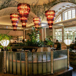 Four amber glass chandeliers glow above people, blurred in motion, who walk between booth table settings, plants and lamps.