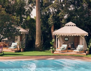 Two private relaxation tents with decks and lounge seating, nestle among garden trees near a corner of the shimmering pool.