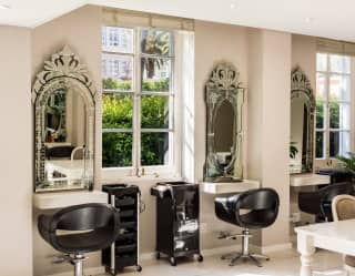 View of four hair-cutting stations with black chairs and ornate silver-framed wall mirrors spaced between two large windows.