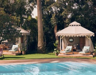 Wind creates brush lines on the blue pool water, overlooked by two cabanas with decks,  sun-loungers and soft pink drapes.