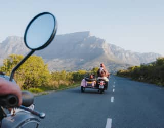 A couple ride a motorbike with a pink sidecar along a road heading towards Table Mountain, seen from a bike riding behind.
