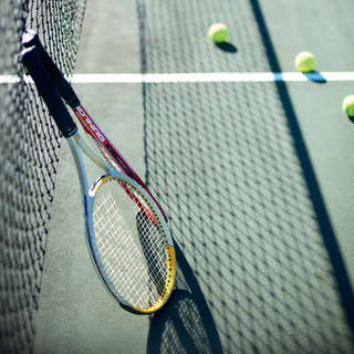 Two tennis rackets lean against the net, which casts a shadow on the green court, where balls have been left during a break.
