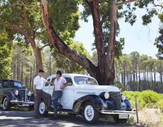 Drivers relaxing against a classic 1930's car on a tree-lined avenue