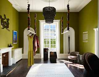 Two turned-wood clothes rails hang from the ceiling above a white rug in an olive room with dark floors and a tall bright window.