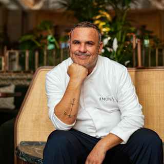 Chef Ángel León smiles to camera as he sits in Amura in a branded white top and dark trousers, resting his chin on his fist.