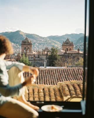 A woman in soft-focus foreground sits on the sill of an open window gazing towards Cusco Cathedral and the mountains beyond.
