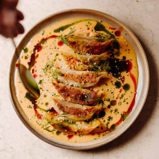 A chef drizzles adobo sauce over a plate of five crisped gyozas, served with a herb garnish, in a close-up, seen from above.