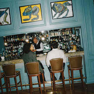 A man and woman sit on two of five high-backed stools in view at the bar as a barman assembles drinks, seen from behind.