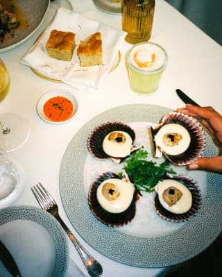 At a dinner table with bread, sauce and a pisco sour, a hand selects one parmesan and truffle scallop from a plate of four.