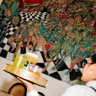 A staff member carries a tray with three cocktails of different hues past a busy, vibrant mural by Mateo Liébana at Tragaluz.