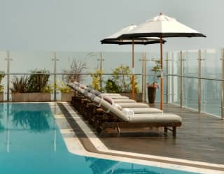 Five white loungers recline under two parasols on the poolside deck, facing views through safety glass, seen over the water.