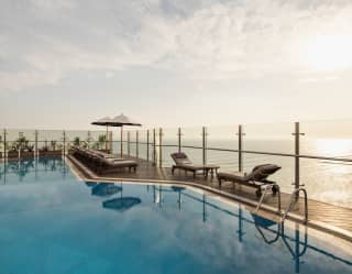 Sun-loungers rest at the glass terrace barrier facing the sunshine glow on the calm ocean, seen across the still blue pool.
