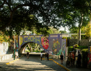 People walk and gather on a cobbled path running beneath the boldly graffitied bridge near Parque Federico Villarreal.