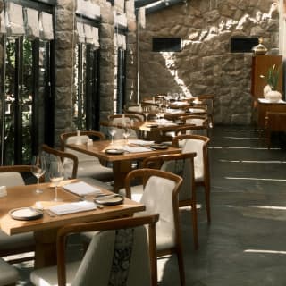 A row of tables along a wall of windows with cutlery and wine glasses, waits for guests beneath the light canopy of Tampu.