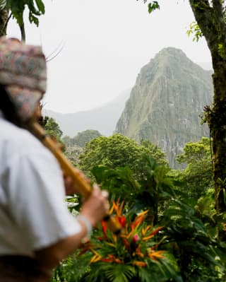 A local shaman wearing a woven hat plays flute facing a view of Huayna Picchu, rising against a cloudy sky.