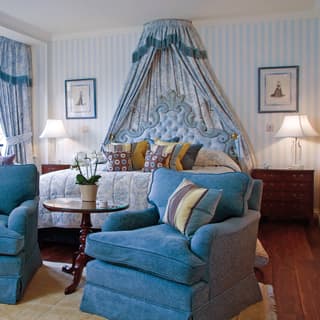 Pale blue floral curtains frame the window and bedhead, matching seat covers and bed quilt beside royal blue armchairs