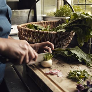 A chef cuts a small white turnip in half. On the chopping board are slivers of radish and a wicker basket overflowing with leaves