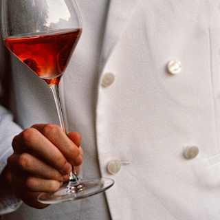 A waiter in a white jacket with pearlescent buttons carries a glass of raspberry-red wine in a close-up dining detail.