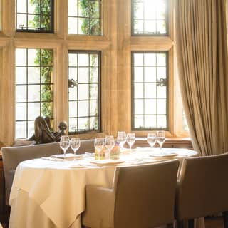 A table set for four occupies the bay of a stone mullioned window nook. A small bronze figure sits on the window sill