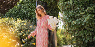 A woman in long summer dress carries a posy of White Asters as she passes a tall bush of yellow flowering St John's Wort