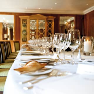 A row of dark wood dining chairs with pale green upholstery sits at a long table, immaculately set for the dinner service