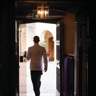 A waiter in white tunic and black trousers carries a tray of drinks in his left hand and he steps out into the garden sunshine