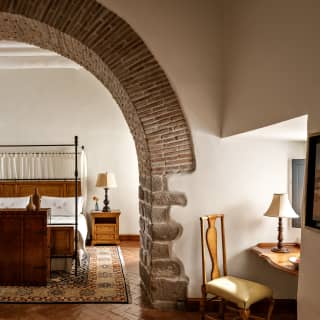 View through a suite's high colonial wall arch with brick edging to an airy bedroom with four-poster bed and luxury rug.