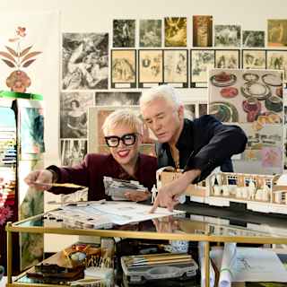 Baz Luhrmann, in black, and Catherine Martin in burgundy with glasses, look through designs in a busy art studio.
