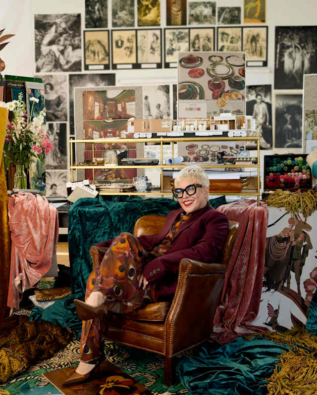 Catherine Martin, in a burgundy jacket over a deep orange-print playsuit, sits in a leather chair among fabrics in a studio.