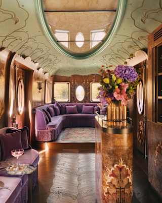 Luxurious vintage train carriage interior with ornate floral decor, plush purple seating, an oval ceiling mirror, flowers on a gold stand, and a built-in bar, all bathed in warm, elegant lighting.