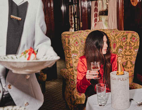 A waiter walks by woman with long dark hair and red satin top as she sits at an Ibis carriage table drinking Veuve Clicquot.