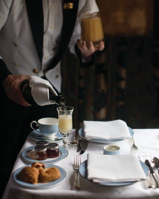 A waiter holds a carafe of orange juice and a bottle of champagne as he pours a glass of Mimosa at a breakfast table for two