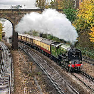 A plume of steam billows out along the length of the cream and burgundy carriages as the train passes below an arched bridge