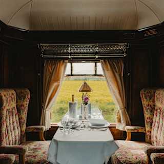 A fields view glows in a window framed by curtains, seen over a dining table flanked by beige and red chairs in the Gwen Car.