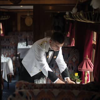 Waiter in a white coat placing silverware on a dining table in a vintage train carriage
