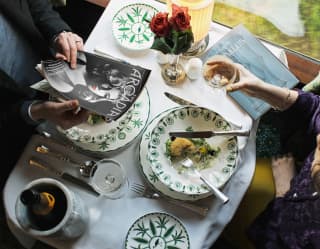 Looking down on a dining table, where a man peruses a magazine and a woman takes a pause mid-way through a starter to drink.