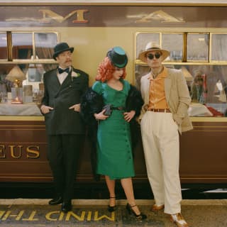 A woman with red curly hair in a green dress and hat leans against the stationed train flanked by two men, all in costume.