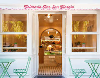 Front view of Gelateria Bar San Giorgia with mint-hued tables either side of a door that opens into a warm, glowing interior.