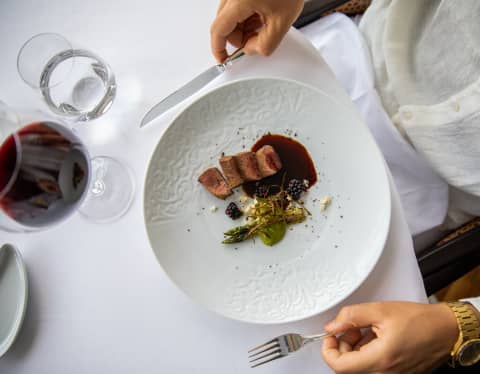 A diner holds a knife and fork by a plate of seared pork and asparagus at a table with red wine and water, seen from above.