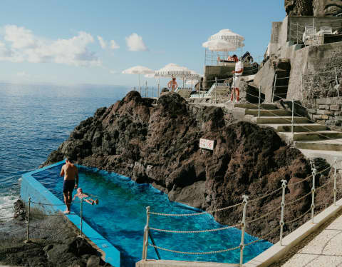 Below the sea platform where loungers and parasols catch sun and spray, two men enjoy the rock pool's proximity to the ocean.