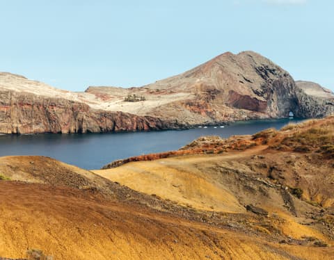 View over a rocky ridge with gold grasses to a deep blue inlet where four small boats are anchored by Ponta de São Lourenço.