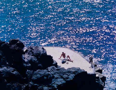 A man lies back on a swimming platform at the base of volcanic rocks, surrounded by glittering azure sea, seen from above.