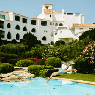 Action shot as a man dives into the still blue pool surrounded by lush gardens and the white Romazzino complex.