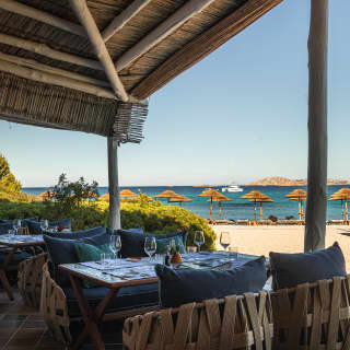 Latticed wicker cocoon chairs with blue cushions provide luxurious casual seating at a Entù e Mari table by the sunny beach.