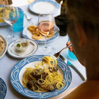 View over a diner's shoulder as he lifts a parcel of spaghetti alle vongole from a blue, fish-patterned plate, at a dinner table.