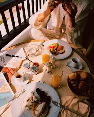 A guest enjoys a balcony breakfast at a sunny table with coffee, orange juice, toast, pastries and fruit, seen from above.