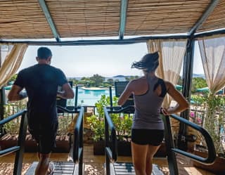 A man and a woman work out side by side on treadmills overlooking the pool in the shaded gym terrace, seen from behind.