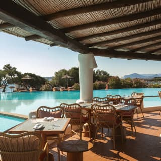 Tables with cane wingback chairs sit in half-sun on the covered pool bar terrace with views over swimming pools and ocean.
